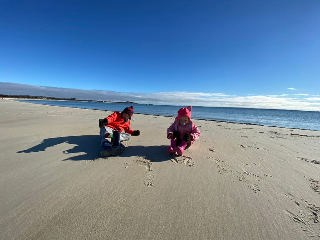 Two kids in winter coats sitting on the sand on a bright blue-sky day, the bay flat behind them.